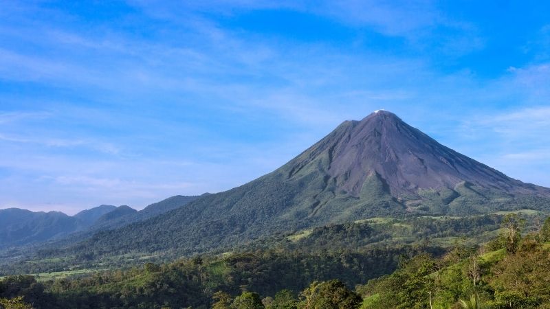 A conical volcano, one of the stunning volcanoes in Costa Rica, rises above lush green forest with a clear blue sky in the background.